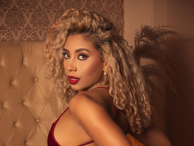 LilithWalker with curly blonde hair, red lipstick, and a red bra, looking over her shoulder, showing her tits, standing in front of a tufted beige headboard.