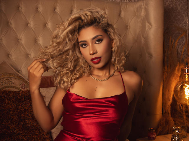 LilithWalker with curly blonde hair, red satin dress, gold chain, and red lipstick, holding her hair, showing her tits, sitting on a bed, looking sexy.