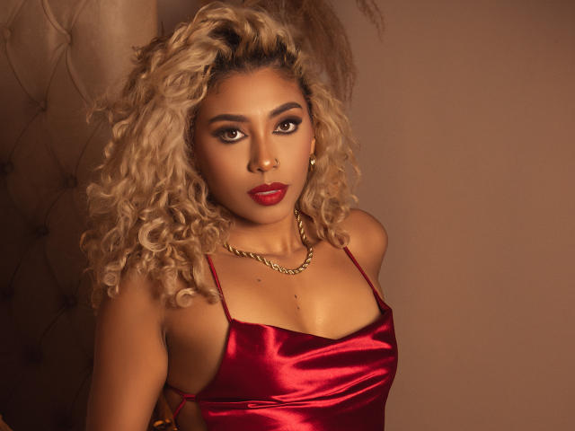 LilithWalker with curly blonde hair, red satin dress, gold chain, red lipstick, and confident gaze, showcasing her boobs and sexy, sultry look in a warm, brown-lit setting.