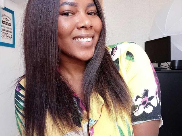 Macarena, avec de longs cheveux noirs, souriante, portant une chemise jaune à motifs floraux, a une peau brun clair et montre des dents blanches. Ses seins ne sont pas visibles sur cette photo..