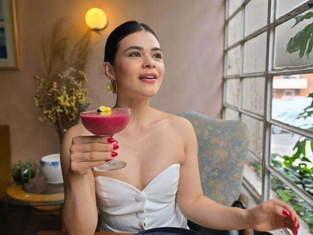 PaulinaValley with dark hair, white strapless dress, holding pink cocktail with lemon slice, red nails, gold earrings, sitting by window, smiling, soft light, indoors.