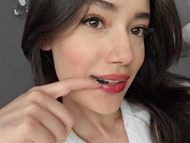 MariaLoft tiene ojos marrón oscuro, lápiz labial rojo y está mordiendo su dedo brillante con una ligera sonrisa. Tiene el pelo largo y castaño, una camisa blanca y un piercing en la nariz..