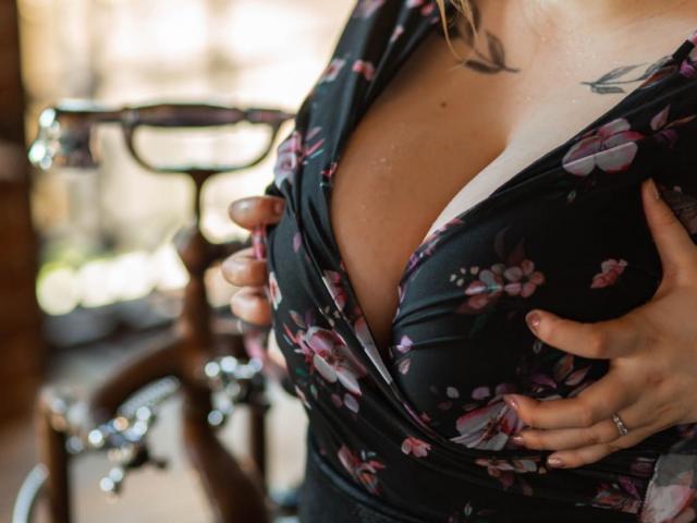 Le mani di SonyaXWilson stringono il suo abito nero con fiori, mettendo in mostra il suo seno prosperoso e il reggiseno bianco. Un piccolo tatuaggio di un uccello è visibile sul suo petto. Le sue dita portano un anello da matrimonio..