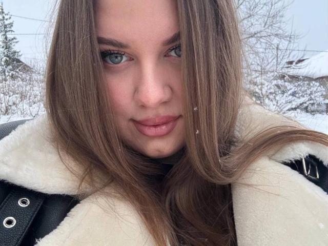 SonyaXWilson has long brown hair, green eyes, and pink lips. She's wearing a white fur-lined coat, looking directly at the camera with a slight smile. Snow is on her face.