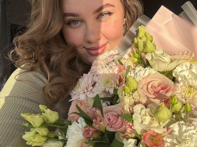 SonyaXWilson, con i capelli biondi ricci, gli occhi verdi e il rossetto rosa, tiene un bouquet di fiori rosa, bianchi e verdi, sorridendo alla fotocamera, con la luce del sole sul viso..