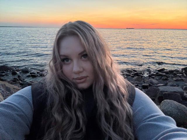 SonyaXWilson, con cabello rubio ondulado, vestida con un chaleco negro sobre un suéter gris, de pie junto a una costa rocosa durante el atardecer, con agua en el fondo, una sonrisa sutil, luz suave de la tarde..