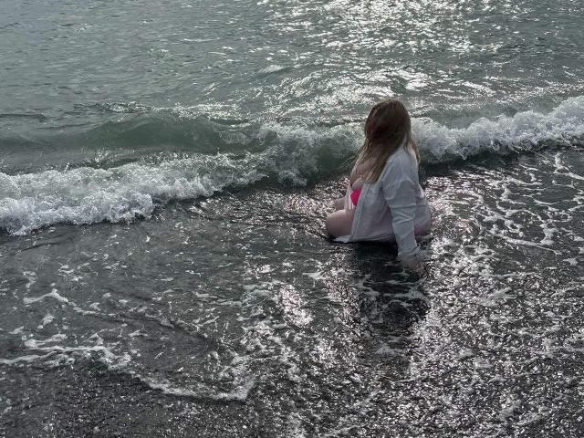 SonyaXWilson sits in shallow water with small waves, wearing a white shirt and pink bikini bottom, her back to the camera, wet blonde hair.