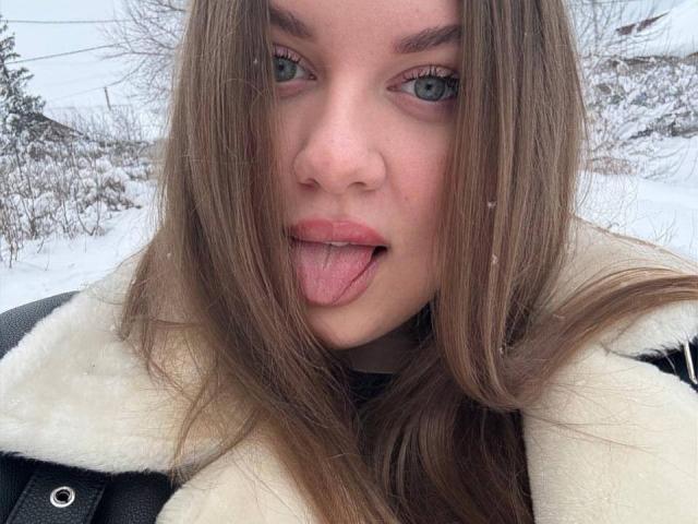 SonyaXWilson has long brown hair, green eyes, and is sticking out her tongue with pink lips. She's wearing a black coat with a white fur collar in a snowy setting.