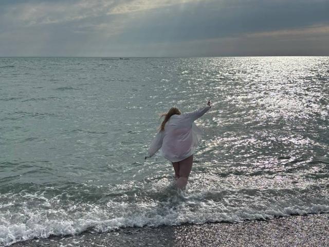 SonyaXWilson con un vestido blanco, de pie en aguas poco profundas con los brazos extendidos, mirando hacia el océano, de espaldas a la cámara, con agua salpicando alrededor de sus piernas, cielo nublado, agua reflectante..