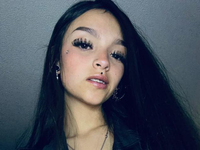 MarietteJones has long black hair, full eyebrows, large eyelashes, a nose piercing, a lip piercing, and a septum piercing, wearing dark earrings and a black top.