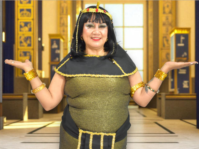MatureYayita with black hair, gold and black costume, gold headpiece, gold bracelets, arms out, smiling, tits visible, standing in a sunlit room with Egyptian-style decor.