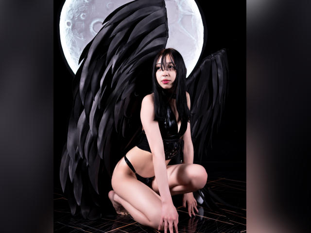 MillaHottLove69 has black wings, pale skin, and long black hair. She's kneeling in black lingerie, showing her ass, with a full moon behind her. She's looking at the camera.
