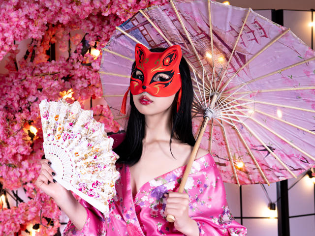 MillaHottLove69 with red mask, pink floral kimono, holding pink fan, and pink parasol, standing in front of pink cherry blossoms, showing off her tits and cute face.