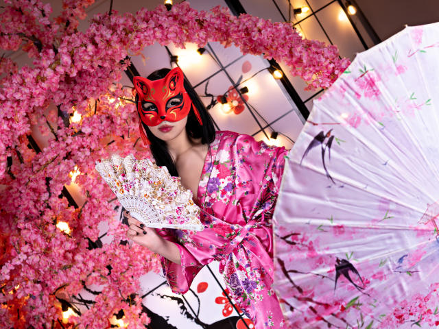 MillaHottLove69 in a pink floral kimono, red mask, holding sparkly fan, pink umbrella, surrounded by pink cherry blossoms, with string lights in background.