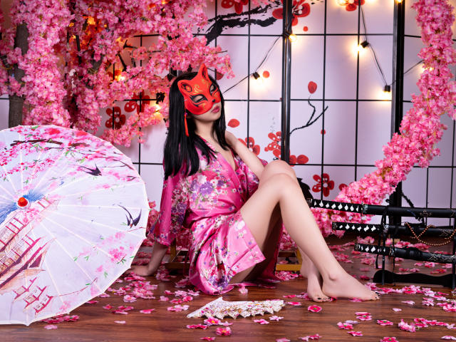 MillaHottLove69 in un kimono floreale rosa, con una maschera rossa, capelli neri, gambe scoperte, seduta su un pavimento di legno, con fiori di ciliegio, un ombrello rosa, una spada samurai, petali, luci calde..