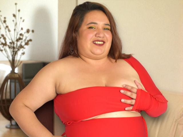 KhloeThompson is a plus-sized woman with brown hair, smiling, wearing a red one-shoulder dress and matching fingerless glove, covering her tits with one hand.