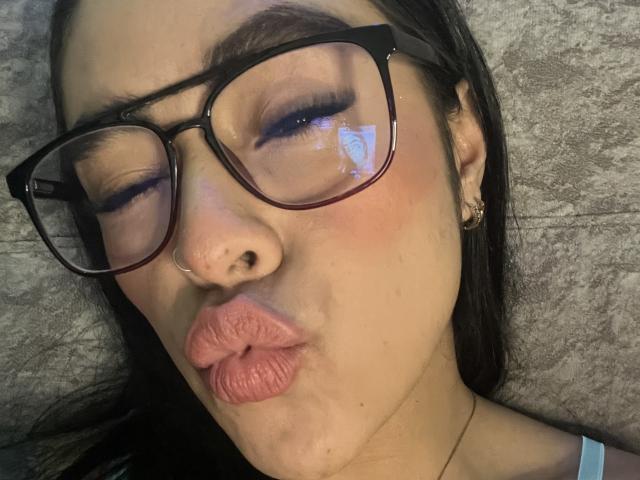 StellaBeckker, con labios rosados y voluminosos, usando gafas negras gruesas, un piercing en la nariz y aretes, acostada con los ojos cerrados y un reflejo en sus gafas..