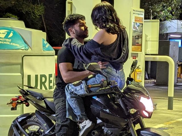 JadeSamuel, with curly black hair and a black off-shoulder top, sits on a black motorcycle, straddling a muscular man with dark curly hair, wearing a black t-shirt.
