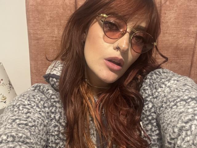 JaneRoa with long red hair, wearing round glasses and a gray knitted sweater, has pink lipstick and a nose ring, looking at the camera with slightly parted lips.