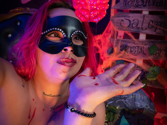 AlanaRoth with red hair, black mask, and nose ring, showing bloody hand, red lips, and beaded bracelet, wearing nothing, under pink light, with a pierced nose and tattoos.