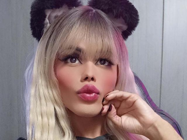 HottAntonella with blonde hair, wearing fuzzy black bear ears, pouty pink lips, and dark nail polish, touching her chin, looking up with sultry eyes.