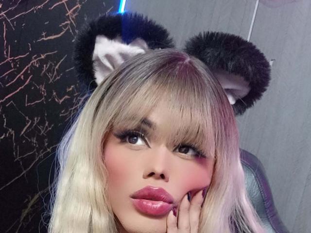 HottAntonella with blonde hair, cat ears, and pink lipstick, looking at the camera with her hand on her cheek. Her tits are small, and she's wearing cute furry ears.