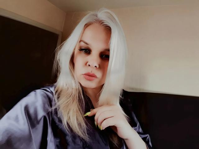 LeilaBlossom with platinum blonde hair, wearing a silky black robe, green nails, and serious expression, holding her chin with her right hand, looking directly at the camera.