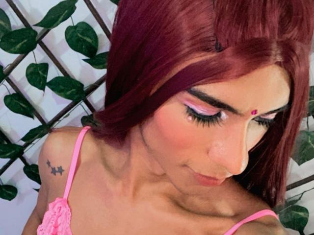 DulceeHott with red hair, pink eyelids, and a pink lace top, looking down with a small star tattoo on her right shoulder. Her tits are visible, and she's cute.