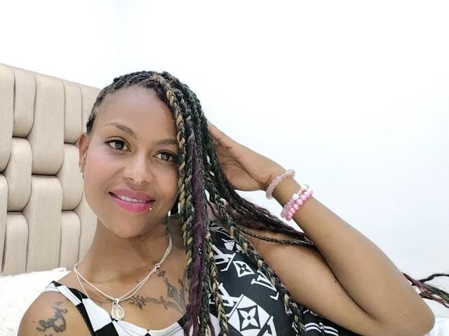 CloeCol has dark skin, braided hair with blonde highlights, pink lipstick, tattoos, a black-and-white patterned top, and a pink bracelet, smiling with a hand on her head.