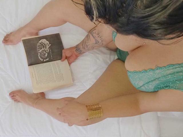 MariGonz's tits in green lace, tattooed arm, gold ankle bracelet, reading a book on white bed, legs spread, brunette hair, tan skin, focused on the open book.