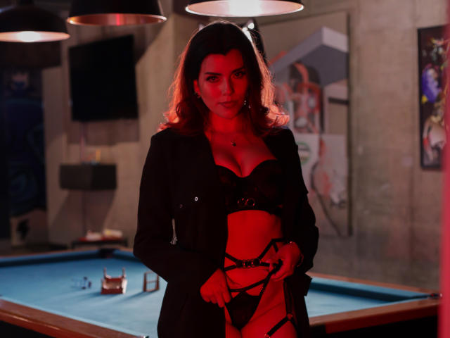 ZoeeMar with red-tinted skin, black lingerie, and strappy red-pink panties, holding a leash, standing in a dimly-lit room with a pool table and hanging lights.