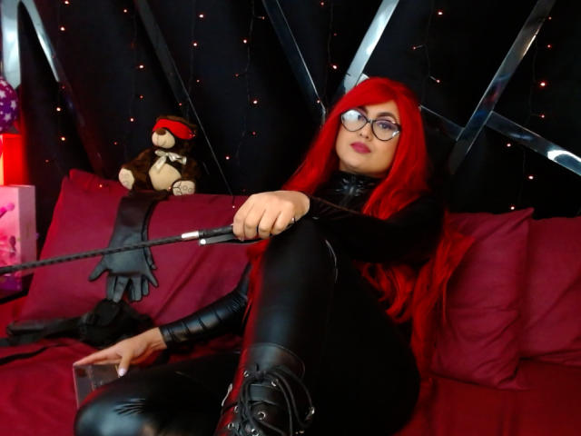 ScarlettRogers with long red hair, glasses, black latex outfit, holding a whip, sitting on red pillows, black boots, stuffed monkey with red hat on left.