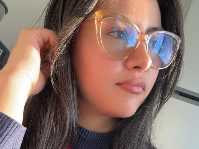 SamaraBetsy, com pele morena, cabelo castanho escuro e óculos de grau, exibindo seus lábios carnudos, vestindo um suéter roxo de malha canelada e tocando sua cabeça..