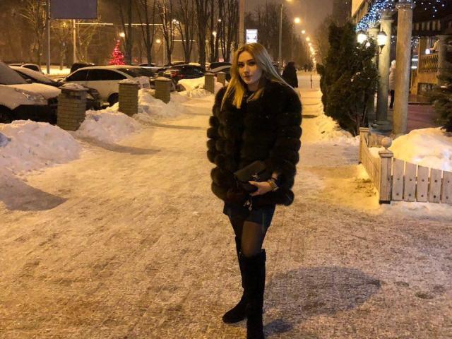CaseyMeli, con cabello rubio largo, vestida con un abrigo de piel negro, una minifalda negra, medias negras y botas negras, de pie en una calle nevada e iluminada por la noche..