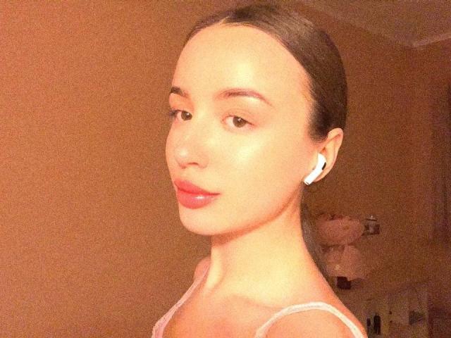 LeslieBer with slicked-back brown hair, wearing white earbuds, pink lipstick, and a pink top, showing off smooth, shiny skin and a cute, subtle smile.