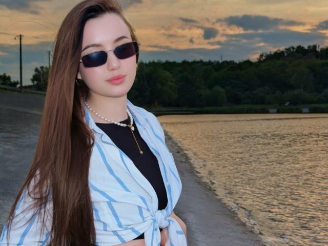 LeslieBer, avec de longs cheveux bruns, portant des lunettes de soleil, un collier de perles et une chemise bleue rayée nouée devant, se tient au bord d'une rivière au coucher du soleil..