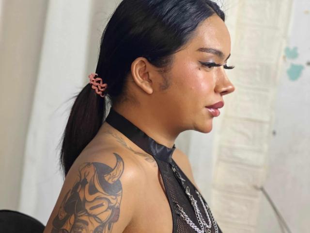 AmaraFoxy with black poneytail, pink scrunchie, black mesh top, shiny lips, tattooed shoulder, chain necklace, looking to the side, soft light, smooth skin, slight smile.