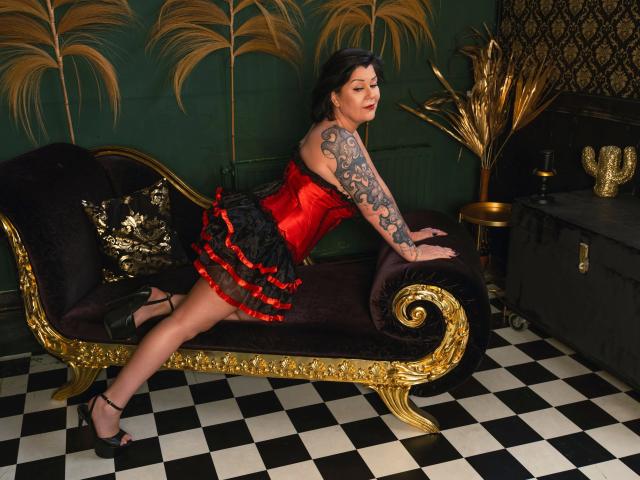 FireFox80-hot with tattoos, red and black corset, ruffled skirt, black heels, leaning on gold and black chaise, checkered floor, dark green wall, gold palm leaves, and black chest.