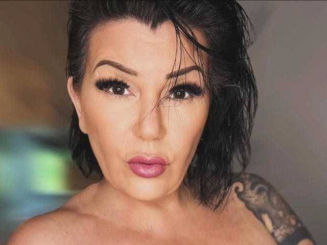 FireFox80-hot has wet black hair, thick eyebrows, long eyelashes, and pink lipstick. She's staring into the camera with a serious expression, showing a tattoo on her right shoulder.