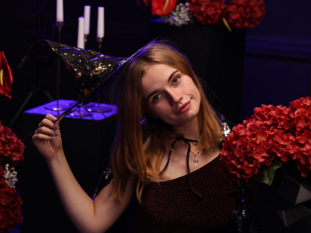 MargaritaKis with blonde hair, wearing a sparkly dark red dress, holding a black glittery party hat, surrounded by red hydrangeas, and white candles in the background.