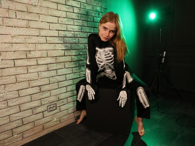 MargaritaKis sits on a black cube, wearing a skeleton costume, her bare feet on dark tile. Green light casts a glow on her, with her long blonde hair.