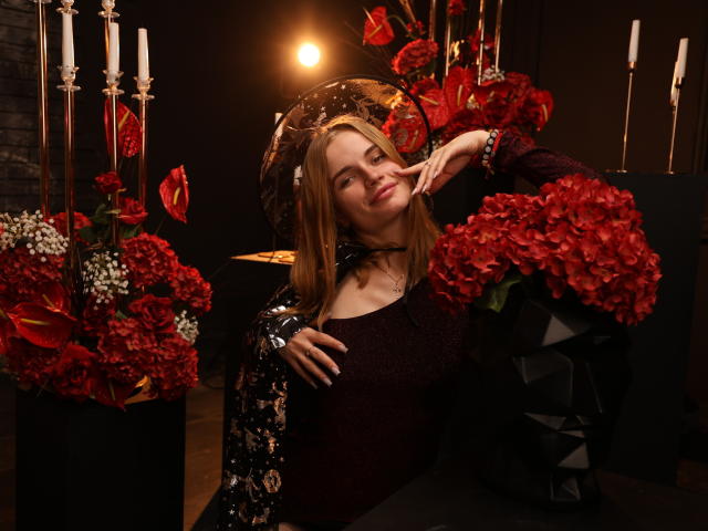 MargaritaKis with red roses, black floral jacket, and dark dress, smiling, hand on cheek, surrounded by red and white flowers, black vase, and white candles.