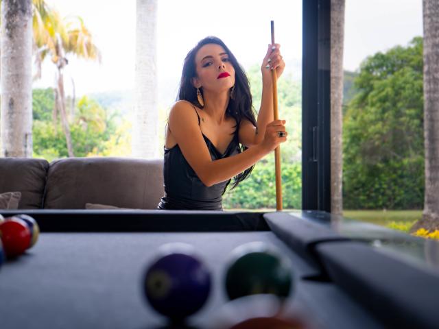 EvelinLuna with black hair and red lipstick, wearing a black tank top, holding a pool cue, standing over a pool table with colorful balls in the foreground.