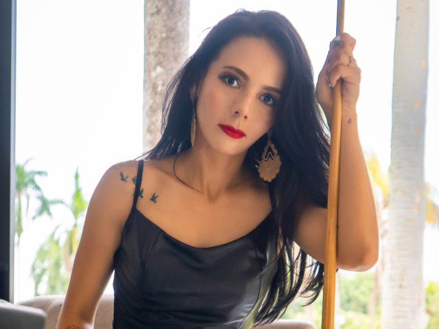 EvelinLuna with long black hair, red lipstick, and black satin dress, holding a wooden pole, showing bird tattoos on her shoulder, wearing large gold earrings, looking at camera.