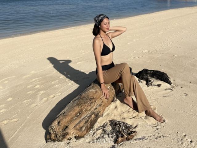 PreciousHorny sits on a rock, wearing a black bikini top, tan pants, and a bandana, with a shadow on the sandy beach, near blue water, looking relaxed.