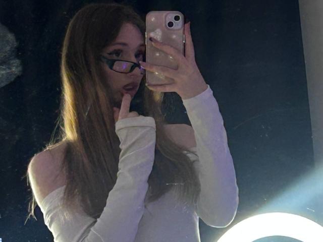 ElfinVibes with long brown hair, glasses, and white off-shoulder top, holding phone, finger in mouth, mirror selfie, ring light on bottom right.