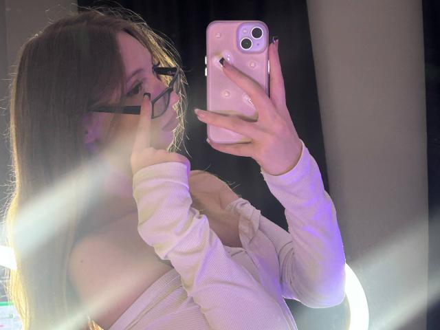 ElfinVibes with glasses, long brown hair, white off-shoulder top, taking a selfie in a mirror, holding a pink phone, visible tits, black nail polish, ring light.