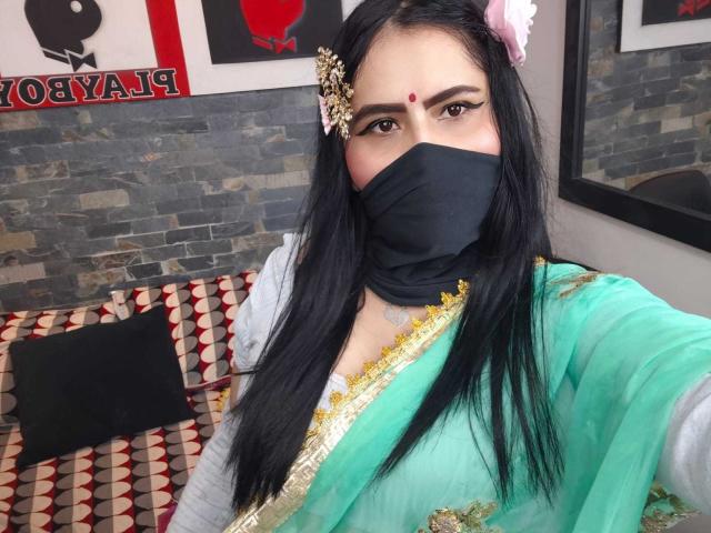 Dalia Dubey mit schwarzer Maske, grünem Sari mit goldener Verzierung, einer Blume im Haar, einem roten Bindi, langen schwarzen Haaren, einem grauen Hemd, sitzend auf einem rot-weißen Sofabezug, mit einer Steinwand im Hintergrund..