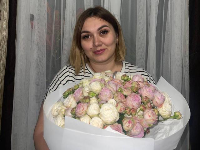 BellSid with shoulder-length brown hair, striped shirt, holding a bouquet of pink and white roses, smiling slightly, looking at the camera, in a well-lit room.