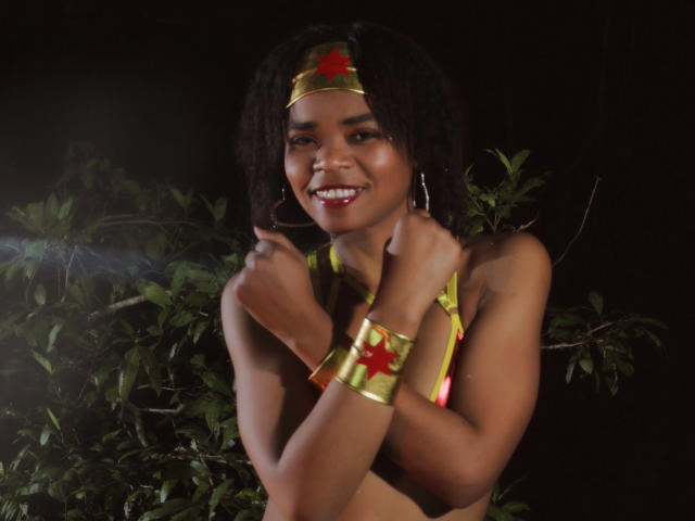 GlettyNoa, com cabelos cacheados escuros, sorrindo, usando uma faixa de cabeça dourada com uma estrela vermelha, um top de biquíni amarelo e pulseiras douradas, mostrando seios pequenos, em uma foto noturna..